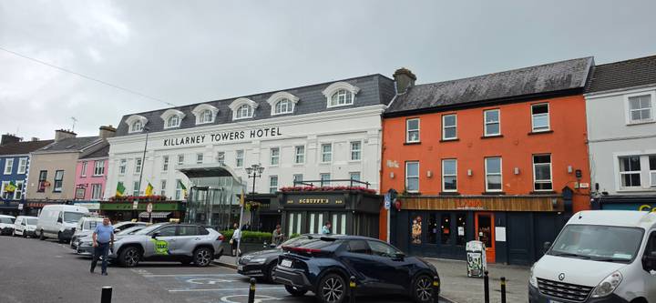 Streetscape of Killarney with Killarney Towers Hotel and shops on a grey day, cars parked in foreground.
