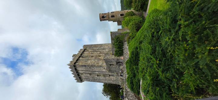 Tall stone Blarney Castle tower rising above green gardens and shrubs under a partly cloudy sky.