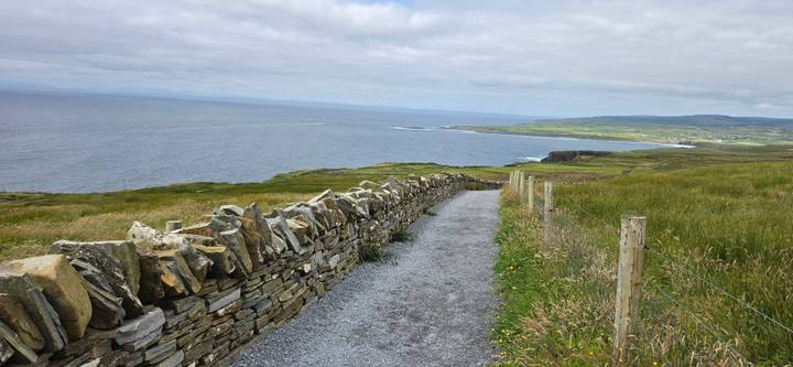 Gravel path lined by stone wall overlooking coastline and distant cliffs under cloudy sky