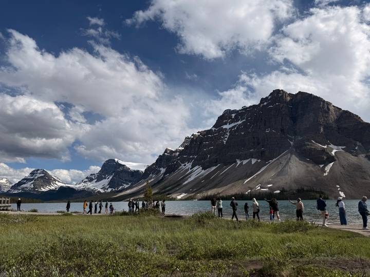 Visitors stand along the shore of Bow Lake with dramatic rocky peaks rising sharply behind.