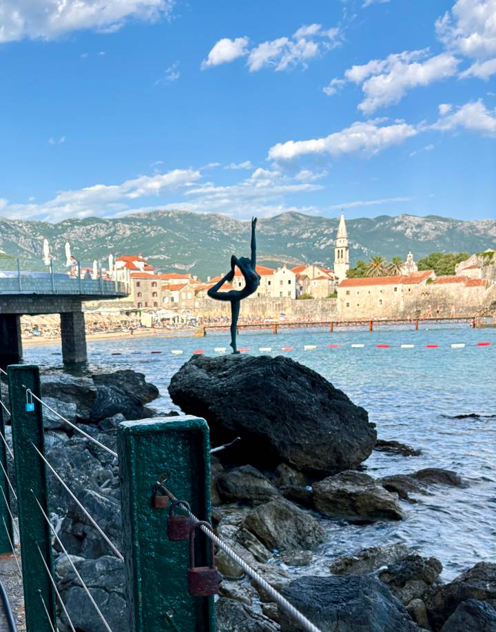 Famous ballerina statue poised on rock with Budva old town and mountains in background.