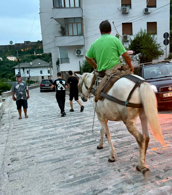 A rider on a white horse passes pedestrians on a cobblestone street with traditional hillside houses in the background.