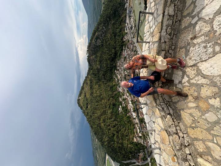 An older couple sits on a stone parapet overlooking a lush hill and the rooftops of a historic town.