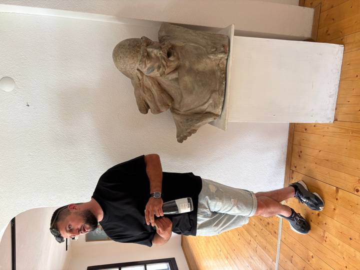 A man holding a wine bottle stands beside an expressive stone sculpture in a bright interior.