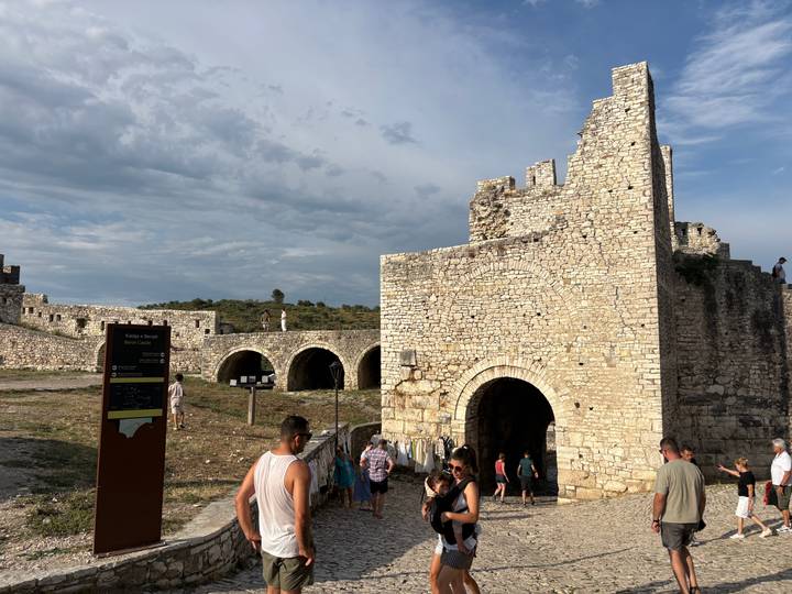 Visitors explore the stone gate and walls of an ancient hilltop fortress under a bright sky.