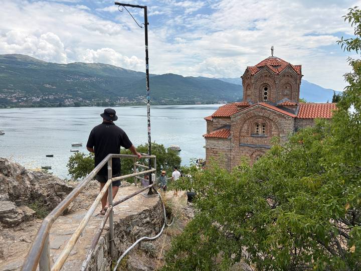 A traveler looks out over Lake Ohrid toward a cliff-top Byzantine church with mountains beyond.