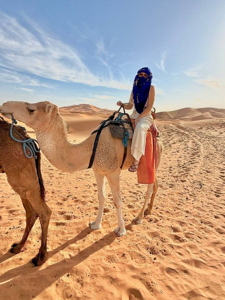 A traveller sits atop a saddled camel amid rolling orange dunes under a pale sky.