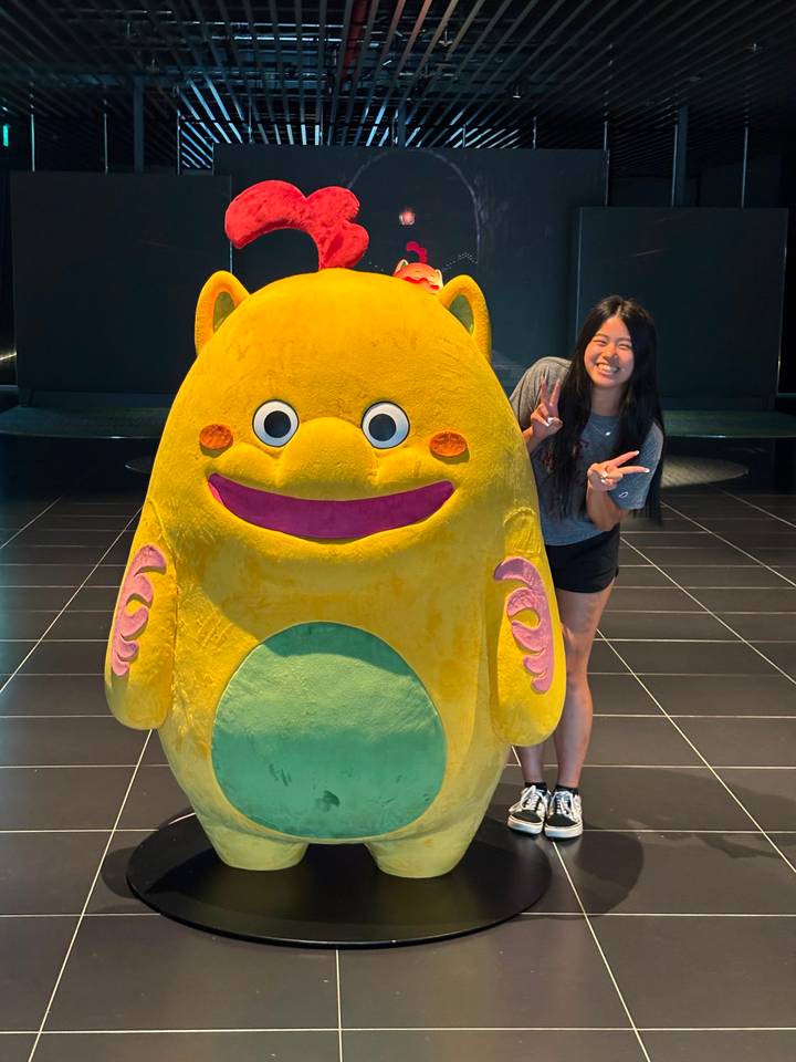 Traveller flashes peace signs next to a large cute yellow character statue in a modern hall