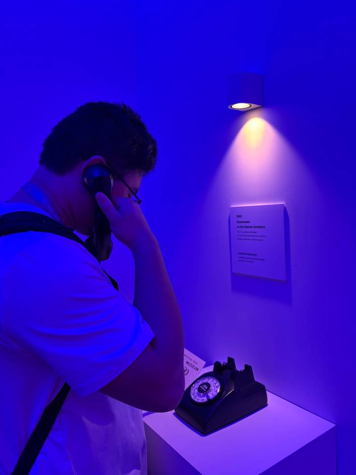Man listens to a vintage telephone handset in a dim blue-lit exhibition space
