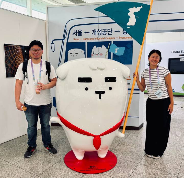 Travellers pose beside a large white mascot statue inside a metro station lobby