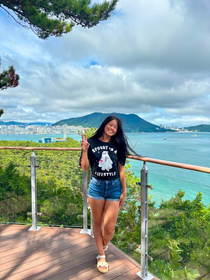 Woman smiles on a wooden deck overlooking turquoise sea and city skyline with forested hills