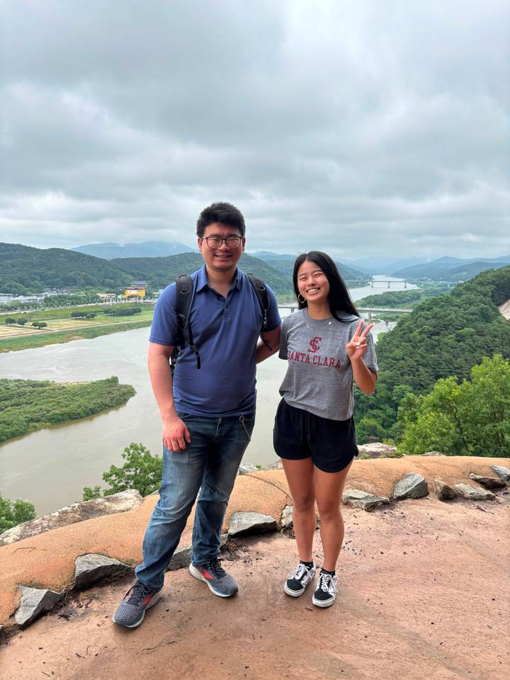 Same couple at the riverside lookout smiling with peace sign against valley backdrop
