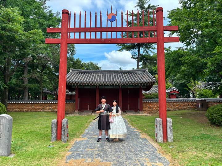 Couple dressed in traditional hanbok stand under a red wooden gate at a historic Korean complex