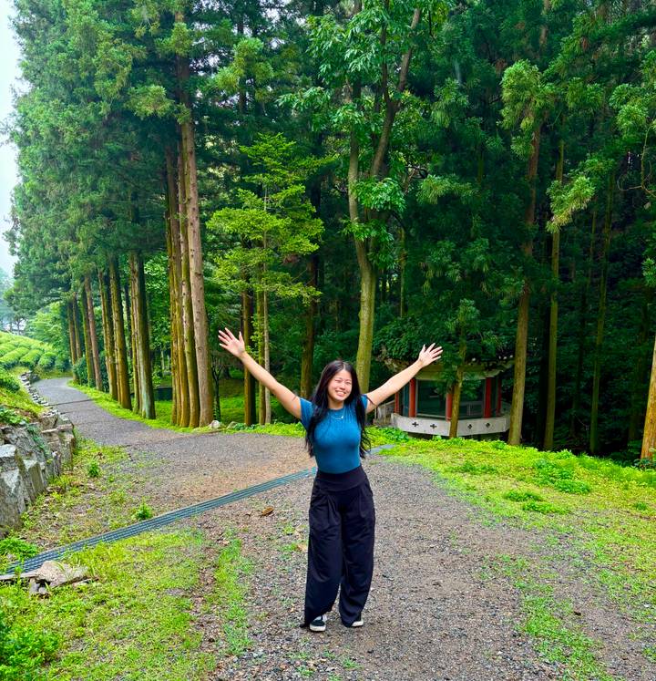 Woman raises her arms happily on a path lined with tall cedar trees in a verdant tea region