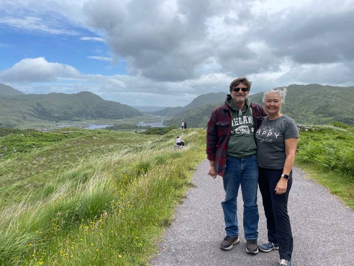 Smiling couple standing on a paved path overlooking green Irish valleys, lakes and mountains under a partly cloudy sky.