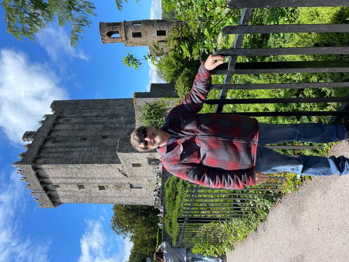 Man posing by an iron fence with the tall stone tower of Blarney Castle rising behind him against a blue sky.