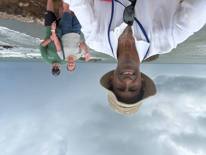 Smiling driver taking a selfie with two travelers on a rocky coastline under stormy skies.