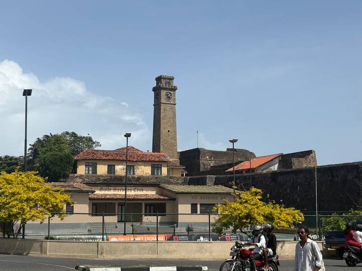 Historic clock tower rising above fort walls with flowering trees and clear blue sky.