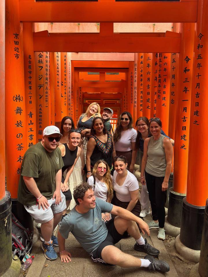 Tour group smiles inside bright orange torii tunnel with Japanese inscriptions.