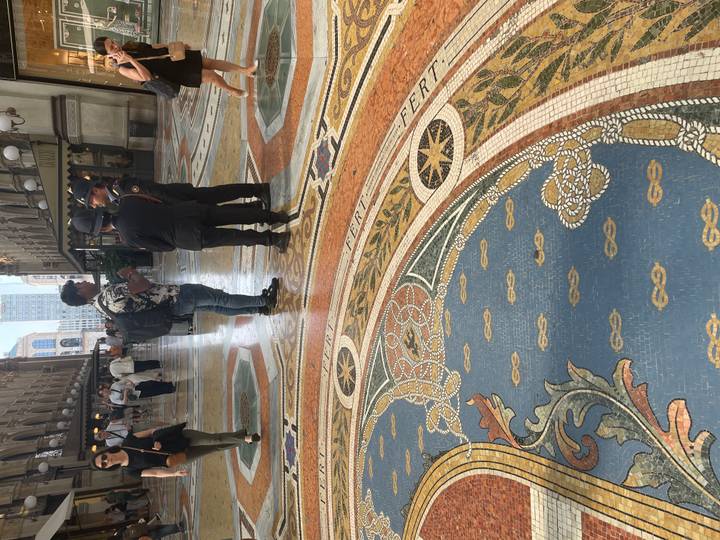 Interior of an elegant shopping arcade with ornate mosaic floor where visitors and uniformed officers converse under glass roof.