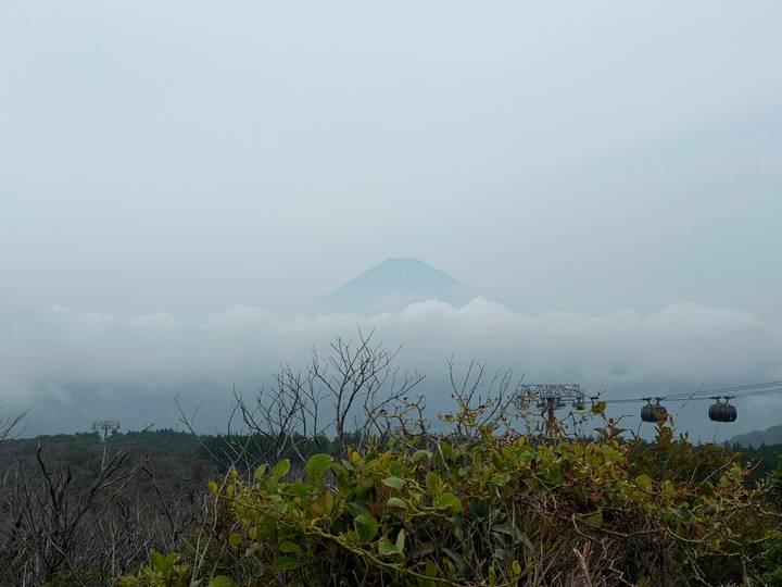 Mount Fuji’s summit barely emerges above a thick layer of clouds on a hazy day, seen behind treetops and cable cars.