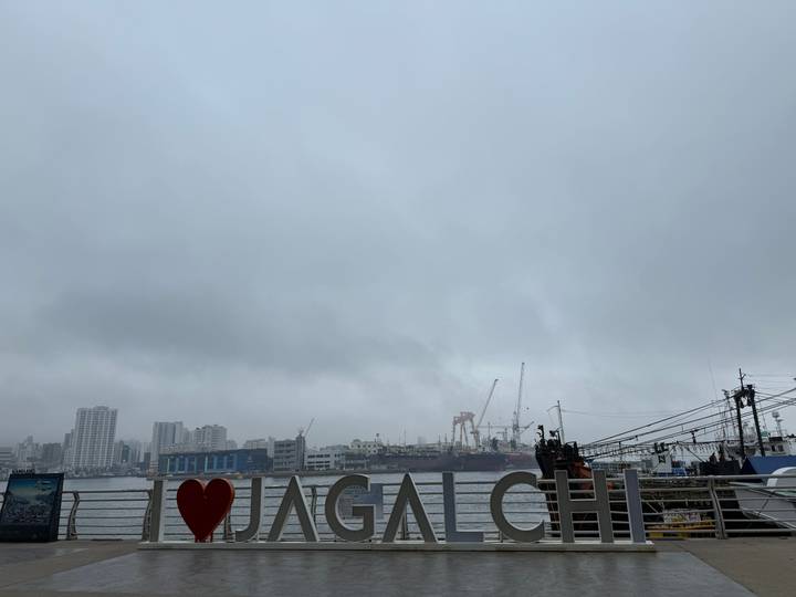 Cloudy waterfront scene showing city buildings, cranes at an industrial port and an “I ❤️” sign in the foreground.
