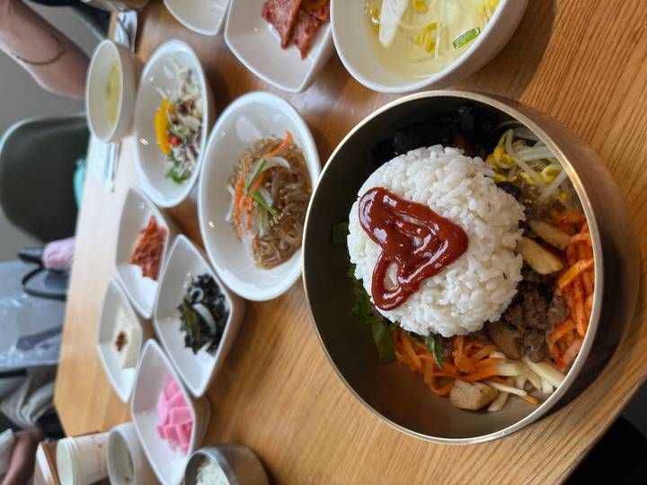 Bowl of bibimbap topped with rice and a heart-shaped dollop of sauce, surrounded by multiple small side-dish plates.
