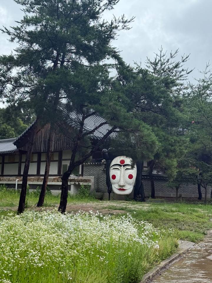 Large traditional Korean mask sculpture set among pine trees with a hanok-style building in the background.