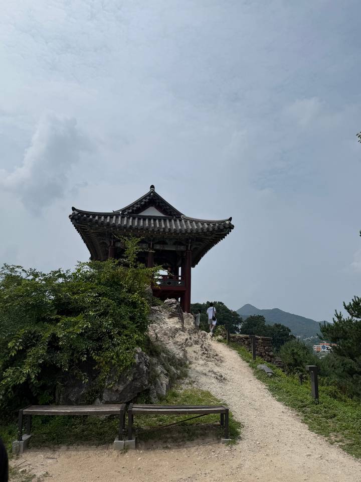 Small pavilion perched on a rocky hilltop with a lone visitor standing below, greenery and hazy mountains behind.