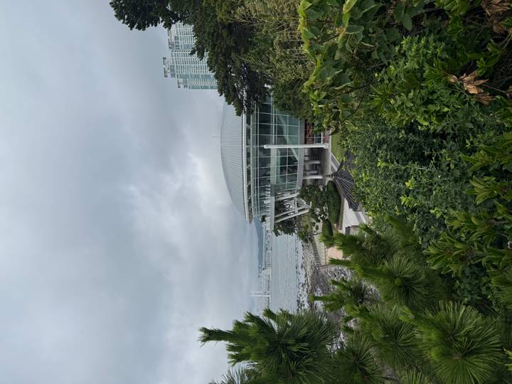 Modern glass-walled rotunda on a coastal cliff with sea, bridge and city skyline partially visible under grey clouds.