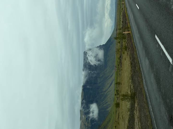 Sheer basalt cliff shrouded in low clouds rising from a grassy plain beside a wet roadway