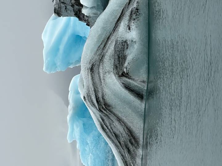 Vertical close-up of a blue and white iceberg with sharp ridges floating in calm grey water
