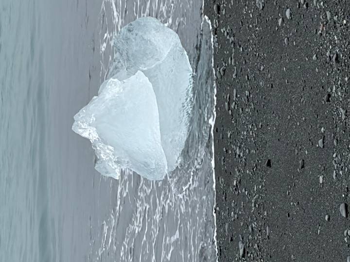 Clear block of glacial ice washed up on black volcanic sand as gentle waves lap the shore