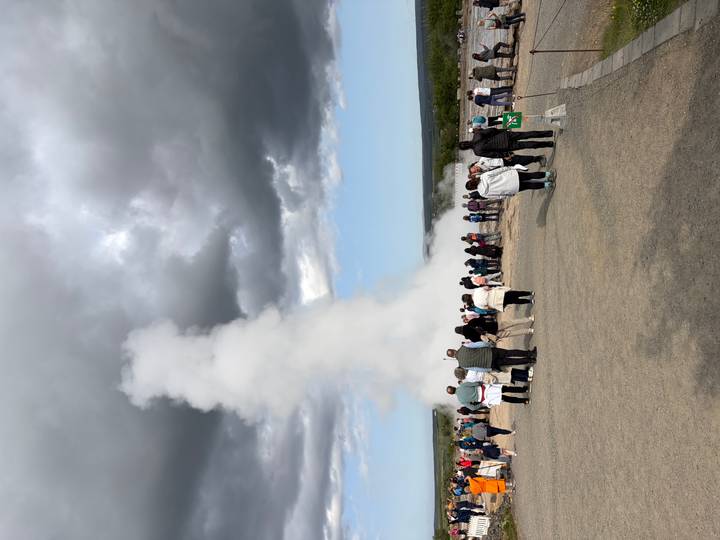 Crowds watch a powerful white geyser erupting against a backdrop of dramatic dark clouds