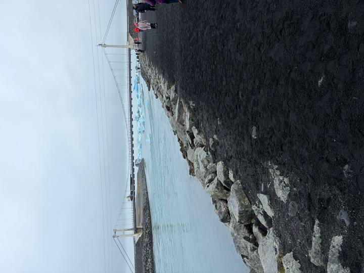 Suspension bridge over a glacial outflow channel with black sand shoreline and distant icebergs