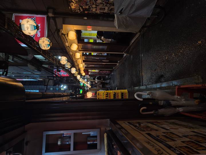 Narrow lantern-lit alley lined with small eateries and bars on a rainy night.