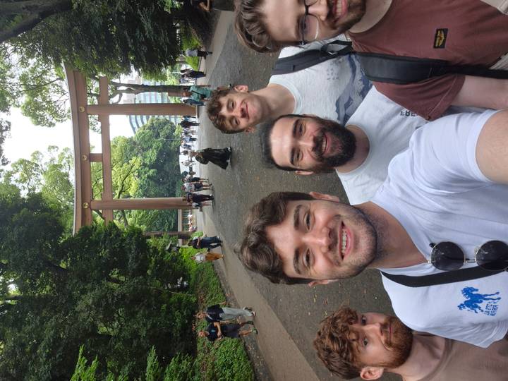 Friends take a selfie beneath a large wooden torii gate in a forested shrine approach.