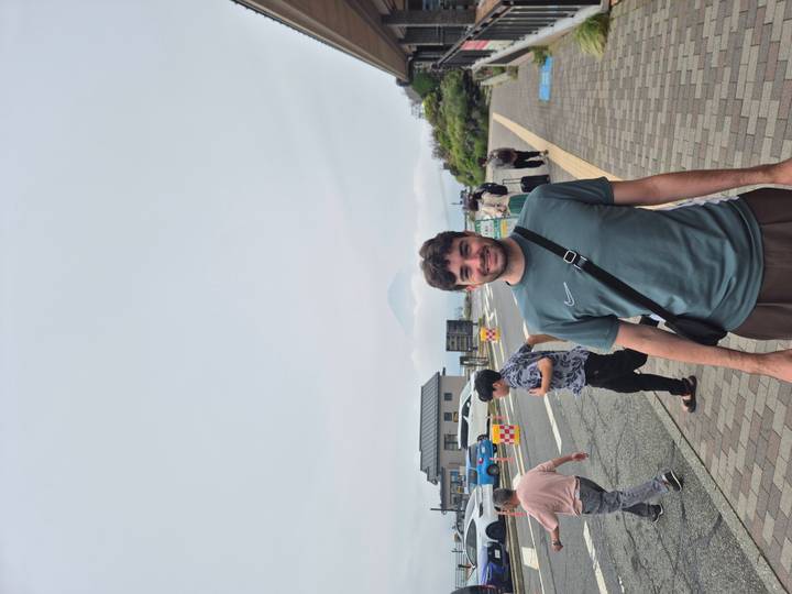Traveller poses at a windy overlook with a faint silhouette of Mount Fuji in the clouds.