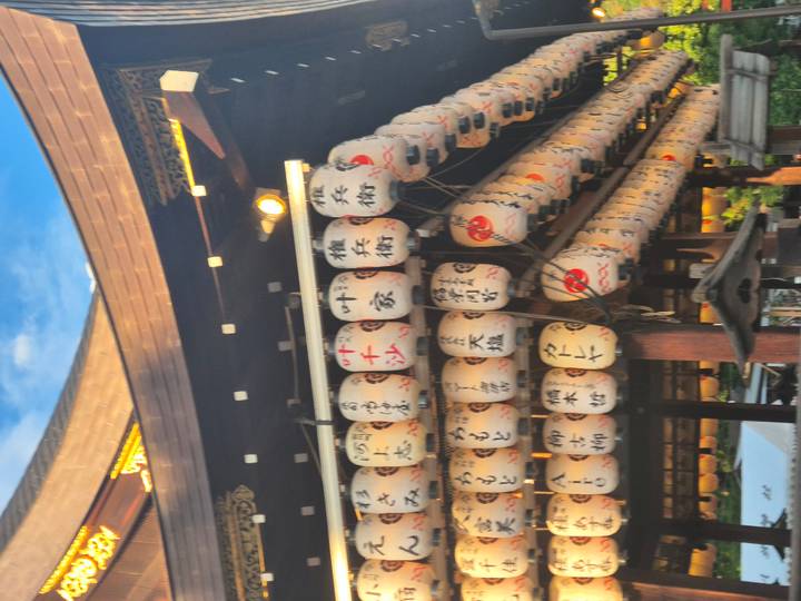 Row of paper lanterns with calligraphy hanging under a wooden shrine roof at dusk.