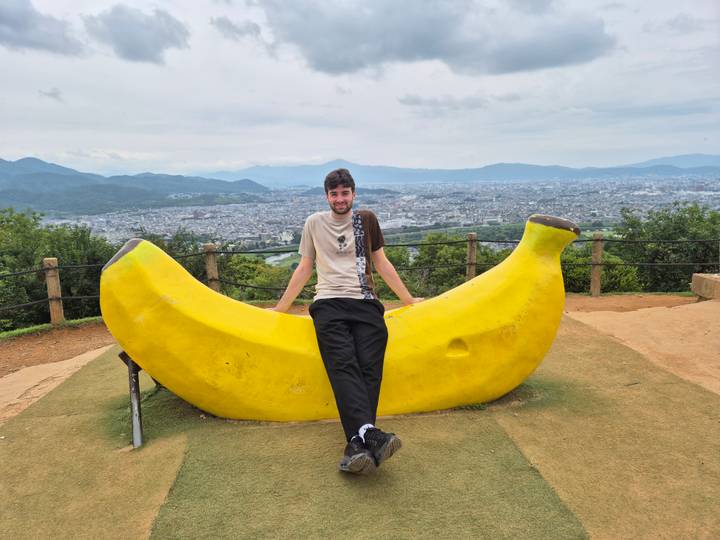 Man sits playfully on a giant yellow banana sculpture overlooking a valley cityscape.