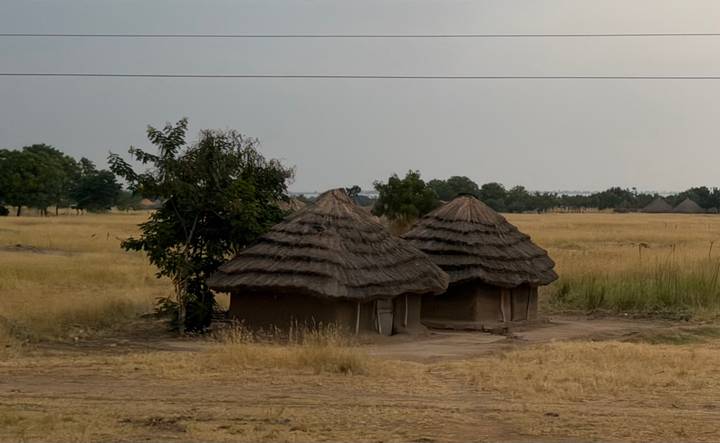 Two traditional thatched huts standing alone in a dry grassland savanna under hazy skies.