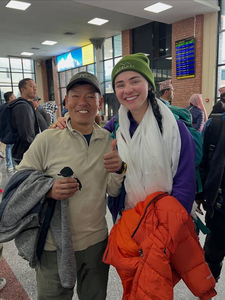 Two travellers smiling and giving thumbs-up inside a terminal, dressed in outdoor gear.