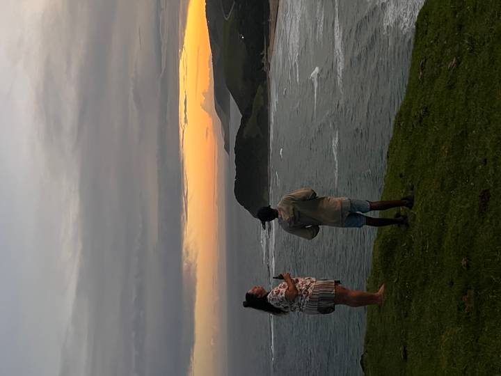 Two travelers standing on grassy cliff watching a colorful sunset over rugged coastline.