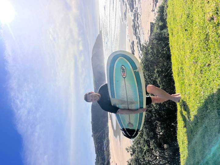 Smiling surfer holding blue board on grassy hill above a sunny beach break.