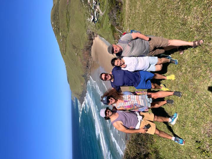 Five hikers posing on a grassy ridge with sweeping ocean bay and cliffs behind them.