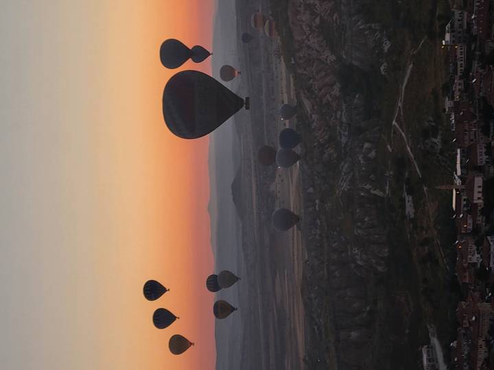 Dozens of hot-air balloons rise over a rocky plateau at dawn, silhouetted against orange sky.