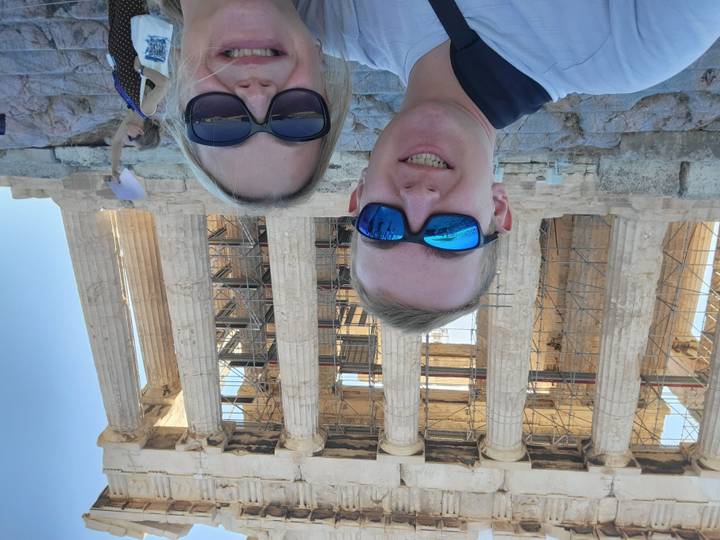 Selfie of two travelers in front of the Parthenon's scaffolding-covered columns in bright sun.