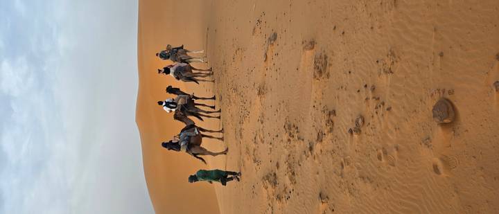 Caravan of tourists riding camels led by a guide across towering sand dunes in the Sahara.