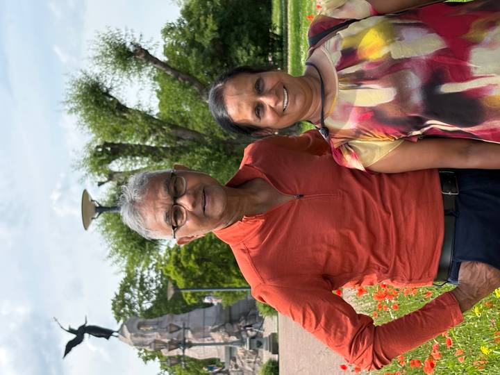 Smiling couple pose in a flower-filled city park beneath leafy trees.