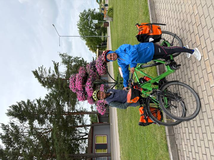 Cycling couple with loaded touring bikes pose beside a bright pink flowering tree in a small town square.